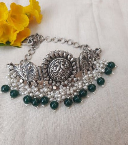 A close-up of the Aadhya Silver Choker Necklace featuring intricate royal motifs, emeralds, and pearls. The silver necklace is adorned with green emerald gemstones and lustrous pearls, creating a regal and elegant design.